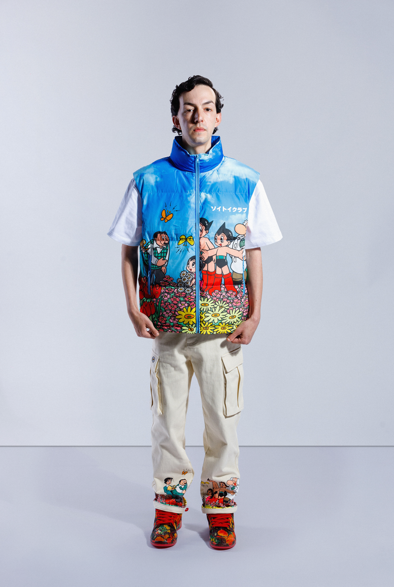 Model wearing the blue Astro Boy Family Portrait Puffer Vest. Features a graphic showing Astro Boy and his family in a field of flowers